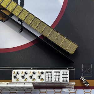 May include: A close-up of a Mahjong set with gold-colored tiles and a black and gold background. The set includes white tiles with black and gold markings, dice, and a gold tile holder. The Luxe Mahjong logo is visible.