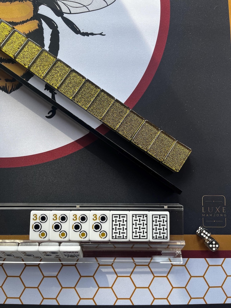 May include: A close-up of a Mahjong set with gold-colored tiles and a black and gold background. The set includes white tiles with black and gold markings, dice, and a gold tile holder. The Luxe Mahjong logo is visible.