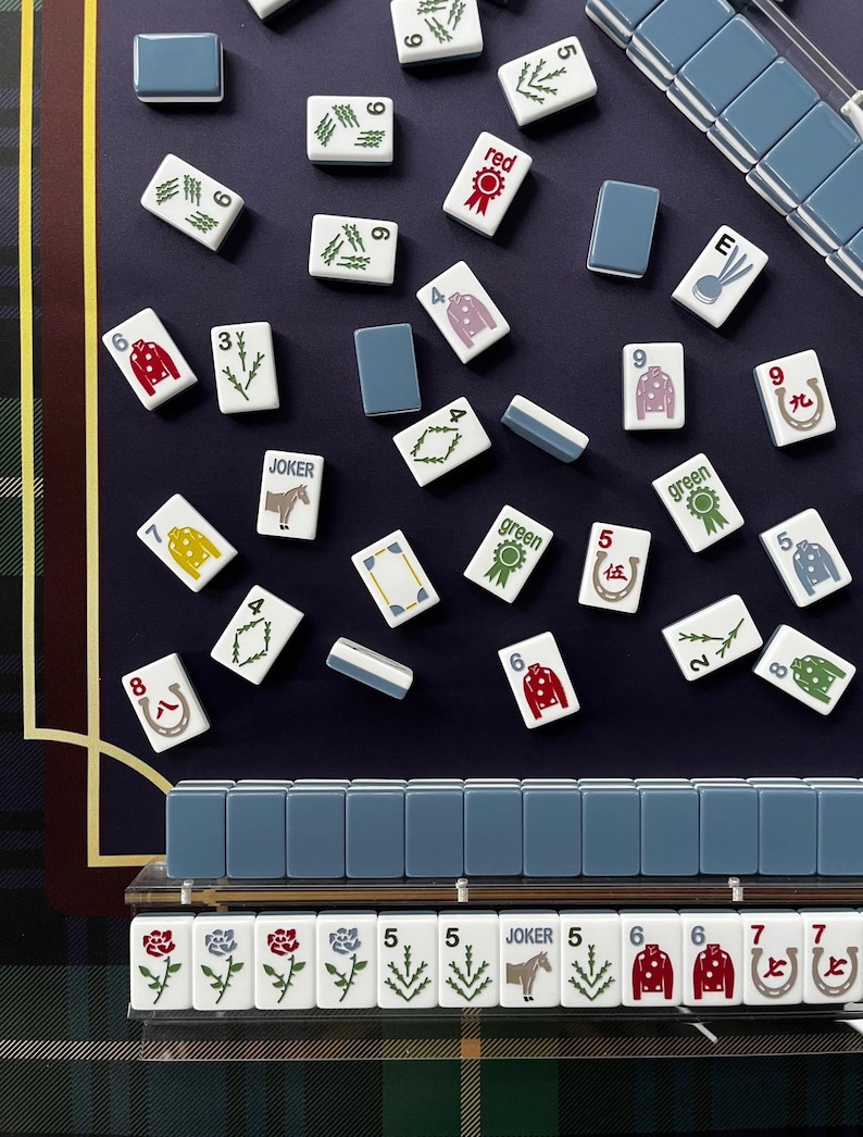 May include: A top-down view of a Mahjong set with white tiles featuring various symbols and numbers. The tiles are scattered on a dark blue surface, with a blue and green plaid pattern in the background. Some tiles have the word "JOKER" on them.
