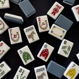 May include: A collection of Mahjong tiles and blue rectangular tiles on a black surface. The Mahjong tiles feature various symbols, numbers, and the word "JOKER". The tiles are white with red, green, and yellow accents.