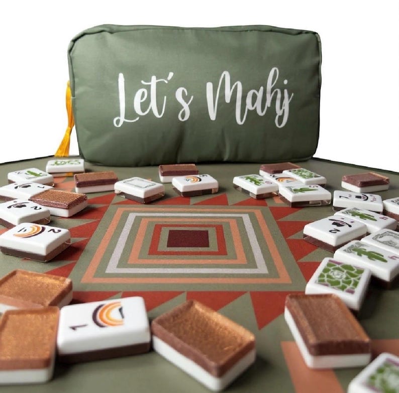 May include: A green Mahjong set with tiles scattered on a patterned mat. The set includes a green carrying case with the words "Let's Mahj" in white script. The tiles are white with brown and colorful designs.