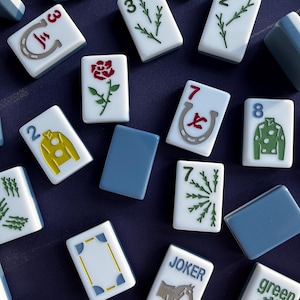 May include: A collection of white and blue Mahjong tiles with various symbols and numbers. The tiles feature images of a rose, a horseshoe, and a horse. Some tiles have the word "JOKER" and "green" printed on them.