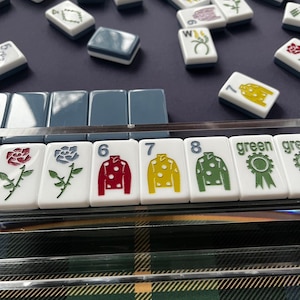 May include: A collection of white Mahjong tiles with various symbols and numbers. Some tiles feature colorful designs like roses and jockey silks in red, yellow, and green. The game is set on a dark blue surface with a green and black plaid pattern visible.