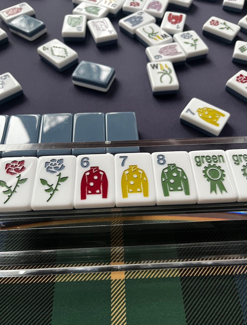May include: A collection of white Mahjong tiles with various symbols and numbers. Some tiles feature colorful designs like roses and jockey silks in red, yellow, and green. The game is set on a dark blue surface with a green and black plaid pattern visible.