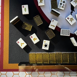 May include: A close-up shot of a Mahjong set with ivory tiles and gold accents. The tiles display various symbols and numbers. The set is arranged on a black surface with a red and gold patterned border. The game set includes a clear acrylic case.