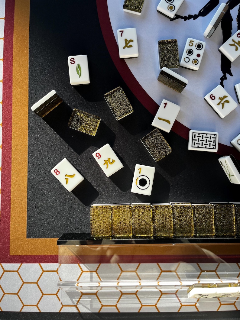 May include: A close-up shot of a Mahjong set with ivory tiles and gold accents. The tiles display various symbols and numbers. The set is arranged on a black surface with a red and gold patterned border. The game set includes a clear acrylic case.