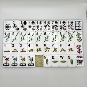 May include: A set of white Mahjong tiles with black, green, red, and gold markings. The tiles feature various symbols, numbers, and Chinese characters. The set includes tiles with the words "NEWS" and "JOKER".