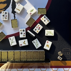 May include: A close-up of a Mahjong set with white tiles featuring various symbols and numbers. Gold-colored dice and decorative gold-colored tile holders are also visible. The game is set on a black and gold patterned surface.