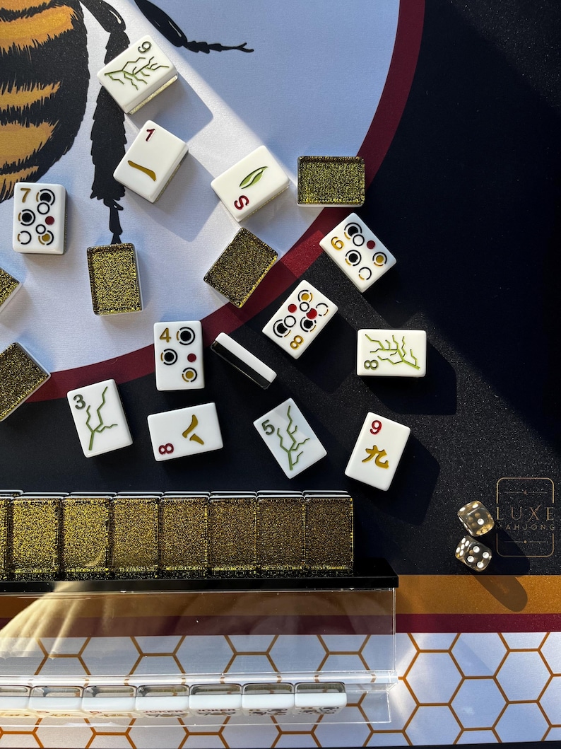 May include: A close-up of a Mahjong set with white tiles featuring various symbols and numbers. Gold-colored dice and decorative gold-colored tile holders are also visible. The game is set on a black and gold patterned surface.