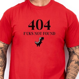 Könnte beinhalten: Rotes T-Shirt mit einem schwarzen Aufdruck, der "404 FCKS NOT FOUND" und ein pixelartiges Dinosaurierbild zeigt.