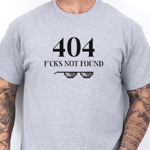 May include: A light grey t-shirt featuring the text "404 F*cks Not Found" in black, with pixelated sunglasses below. The short-sleeved shirt is likely made of a soft fabric.