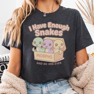 Op de afbeelding: Donkergrijs t-shirt met de tekst "I Have Enough Snakes" en een cartoonafbeelding van drie kleurrijke slangen in een doos. De tekst "MORPHS SAID NO ONE EVER" staat eronder. Het shirt is een casual stijl.