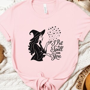 May include: A light pink t-shirt featuring a black graphic of a witch in a hat, conjuring stars and hearts. The text "I Put a Spell on You" is below the witch. The shirt is made of a soft material.