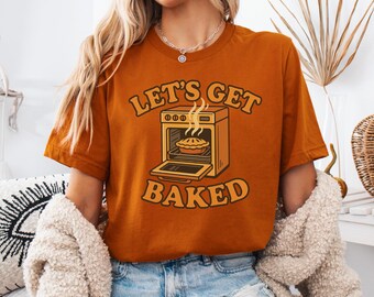 Puede incluir: Camiseta naranja quemado con el texto "Let's Get Baked" sobre un horno de dibujos animados horneando un pastel. La puerta del horno está abierta y sale vapor del pastel. La camiseta es de color liso y tiene mangas cortas.