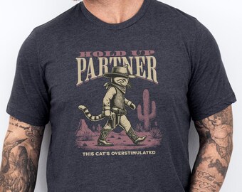 May include: A dark gray t-shirt with a cartoon cat wearing a cowboy hat and holding a gun. The text "Hold Up Partner" is above the cat and the text "This Cat's Overstimulated" is below the cat.