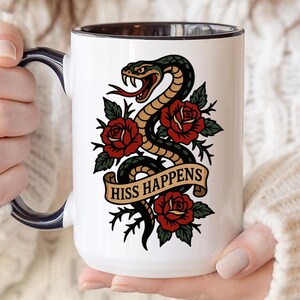 May include: White ceramic mug with a black interior and a navy blue handle. The mug features a tattoo-style design of a green and black snake, red roses, and the words "HISS HAPPENS" on a banner.
