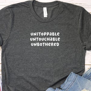 Puede incluir: Una camiseta gris oscuro con el texto "Unstoppable Untouchable Unbothered" impreso en blanco.