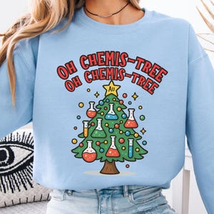 Puede incluir: Sudadera azul claro con un diseño de árbol de Navidad. El árbol está decorado con matraces de laboratorio y el texto "OH CHEMIS-TREE" en rojo y naranja. Una estrella dorada con "Au" se encuentra en la parte superior del árbol.