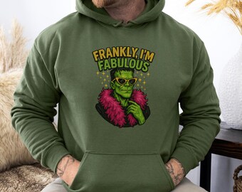 Puede incluir: Sudadera con capucha verde con un gráfico frontal de Frankenstein con gafas de sol y una boa rosa. El texto "FRANKLY, I'M FABULOUS" está encima del gráfico en amarillo y verde. La sudadera tiene un bolsillo delantero y una capucha con cordón.