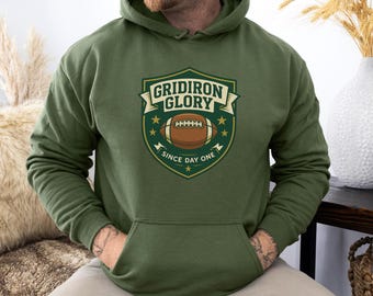 May include: Olive green hoodie with a front pocket and a drawstring hood. The front features a shield-shaped graphic with a football, stars, and the text "GRIDIRON GLORY SINCE DAY ONE" in white and gold.