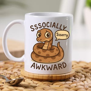 Könnte beinhalten: Weiße Keramik-Tasse mit einem Cartoon-Schlangen-Design. Die braune Schlange hat dunklere Flecken und ist aufgerollt. Der Text auf der Tasse lautet "SSOCIALLY AWKWARD" mit einer Sprechblase mit der Aufschrift "Sssorry".