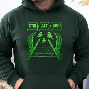 May include: Dark green hoodie with a graphic design featuring a keyboard, skeleton hands, and ghosts. The text "CTRL + ALT + BOO!" is displayed above the keyboard. A fun Halloween-themed design.