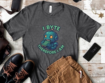 Könnte beinhalten: Dunkelgraues T-Shirt mit einer türkisfarbenen Roboter-Grafik, die eine Platine isst. Der grüne Text lautet "I BYTE THEREFORE I AM". Das Shirt wird mit brauner Hose, schwarzen Schuhen und einem Telefon präsentiert.