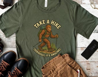 May include: Olive green t-shirt with a Bigfoot graphic hiking with the text "Take a Hike (Seriously, It's Amazing)". The shirt is paired with brown pants and black shoes. A smartphone and other accessories are also visible.