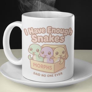 May include: White ceramic mug with the text "I Have Enough Snakes" and a cartoon illustration of three colorful snakes in a box. The mug is on a white saucer, with steam rising from the top.