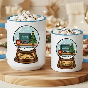 Puede incluir: Dos tazas de cerámica blancas con asas y bordes azules, llenas de malvaviscos. Cada taza presenta un diseño de bola de nieve con un árbol de Navidad, una pantalla de ordenador y el texto "May Your Code Compile."