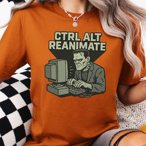 Puede incluir: Una camiseta naranja con el texto "CTRL ALT REANIMATE" y un gráfico del monstruo de Frankenstein usando una computadora. El monstruo está representado en verde y gris, con una expresión seria. La camiseta es de color sólido.