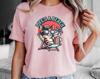 May include: Pink t-shirt featuring a cartoon cat wearing sunglasses and a hat, holding a beer and relaxing in a beach chair. The text "Life's a Beach" is above the cat.
