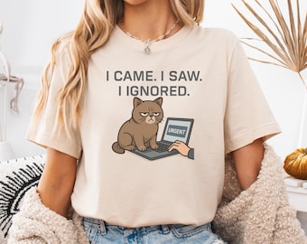 May include: Beige t-shirt with the text "I CAME. I SAW. I IGNORED." A cartoon cat sits in front of a laptop with the word "URGENT" on the screen. The cat has a grumpy expression. The shirt is a casual style.