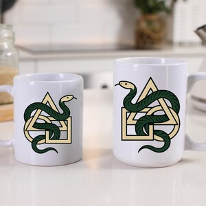 Puede incluir: Dos tazas de cerámica blanca con un diseño de serpiente verde. La serpiente se enrolla alrededor de una forma geométrica con un triángulo, un cuadrado y un círculo. La serpiente tiene una cabeza amarilla y está sobre un fondo blanco.