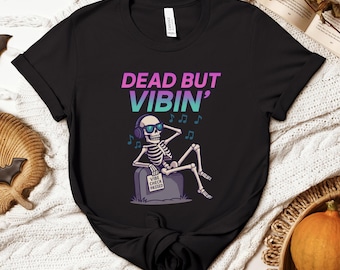 May include: Black t-shirt with the text "DEAD BUT VIBIN'" in a gradient of purple and teal. A cartoon skeleton wearing sunglasses and headphones is relaxing on a tombstone that reads "VIBE CHECK PASSED". Musical notes float around the skeleton.