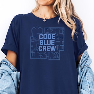 Peut inclure: T-shirt bleu marine avec un motif bleu clair. Le motif comprend le texte "CODE BLUE CREW" et un dessin de type plan avec un stéthoscope. Le t-shirt a des manches retroussées.