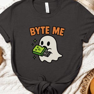 May include: Dark grey t-shirt featuring the orange text "BYTE ME" above a cartoon ghost. The ghost is holding a green and grey game controller. The ghost has a white body and green sparkles.