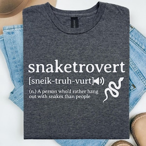 Puede incluir: Una camiseta gris oscura con un gráfico blanco que dice "snaketrovert" y lo define como "una persona que prefiere pasar tiempo con serpientes que con personas". El gráfico también incluye una serpiente y un icono de altavoz.