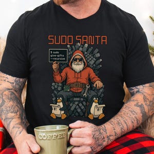 Op de afbeelding: Zwart T-shirt met een afbeelding van de Kerstman in een rode hoodie, zonnebril en witte baard, zittend op een troon van USB-drives. De tekst "SUDO SANTA" staat boven de afbeelding, en een bord leest "$ sudo give-gifts --recursive".