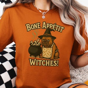 以下が含まれることがあります： ハロウィーンをテーマにしたオレンジ色のTシャツ。魔女の帽子とエプロンを着たブルドッグ、大釜、そして「Bone Appétit Witches!」というテキストが遊び心のあるフォントでデザインされています。