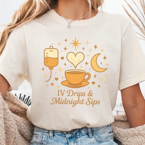 May include: Cream-colored t-shirt featuring a graphic design with an IV drip bag, a heart, a coffee cup, and a crescent moon in shades of orange and gold. The text "IV Drips & Midnight Sips" is below the graphic. The shirt is made of soft material.
