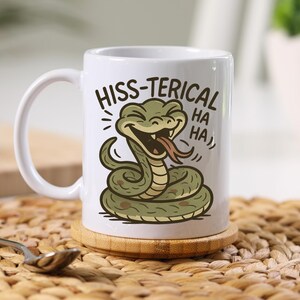 Peut inclure: Mug en céramique blanche avec une illustration de serpent de dessin animé. Le serpent est vert et beige, avec sa langue sortie. Le texte sur la tasse dit "HISS-TERICAL HA HA HA". La tasse est posée sur un dessous de verre en bois.