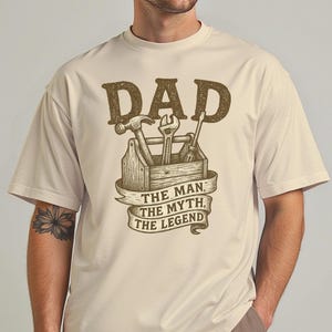 Könnte beinhalten: Ein beiges T-Shirt mit einer braunen Grafik einer Werkzeugkiste mit Werkzeugen und dem Text "DAD THE MAN, THE MYTH, THE LEGEND".