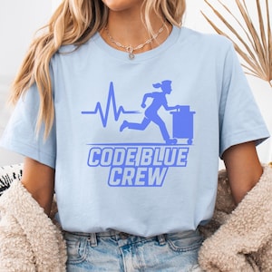 May include: Light blue t-shirt with a graphic featuring a running figure pushing a cart, a heartbeat line, and the text "CODE BLUE CREW" in blue. The shirt is made of a soft material.