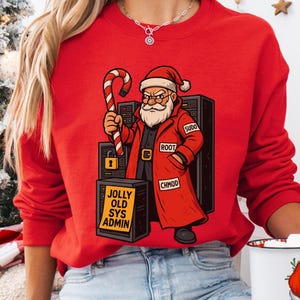 Op de afbeelding: Rode sweatshirt met een cartoon kerstman die een zuurstok vasthoudt. De kerstman draagt een rode jas en hoed. Het ontwerp bevat computer server racks en de tekst "JOLLY OLD SYS ADMIN".