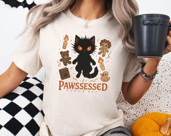 Może przedstawiać: Kremowa koszulka z czarnym kotem o pomarańczowych oczach i napisem "PAWSSESSED". Grafika zawiera ilustracje świec, misia, lalki, książki i gumowej kaczki.