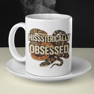 May include: White ceramic mug featuring the text "HISSSSTERICALLY OBSESSED" and snake illustrations in brown, orange, and black. The mug sits on a white saucer, with steam rising from the top.
