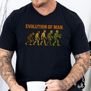 May include: Black t-shirt with the text "EVOLUTION OF MAN" in orange letters. The graphic depicts the evolution of man, transitioning into a zombie. The shirt is a crew neck and has short sleeves.