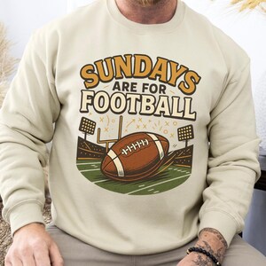 Op de afbeelding: Beige sweatshirt met de tekst "SUNDAYS ARE FOR FOOTBALL" in oranje en wit. De afbeelding toont een voetbal, stadionlichten en een veld.