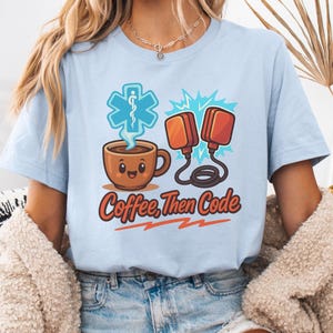 Op de afbeelding: Lichtblauw T-shirt met de tekst "Coffee, Then Code" in rood en oranje. De afbeelding bevat een lachende koffiekop, een medisch symbool en defibrillatorpeddels. Het shirt is gemaakt van een zacht materiaal.